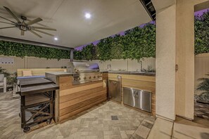 An outdoor kitchen area with a grill, sink, and storage cabinets, surrounded by a patio with a ceiling fan and a wall of greenery. - Breezin': Pool, Spa, Fire Pit, Foosball and more! (Indio)