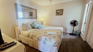 Ein Schlafzimmer mit einem Bett, einem Nachttisch mit Fernbedienung, einem Standventilator und einem Fenster mit Jalousien.