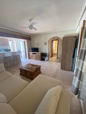 A spacious living area with a sofa, a wooden coffee table, and a television. There is a kitchen area with a refrigerator and a dining table with chairs. An arched doorway leads to another room.