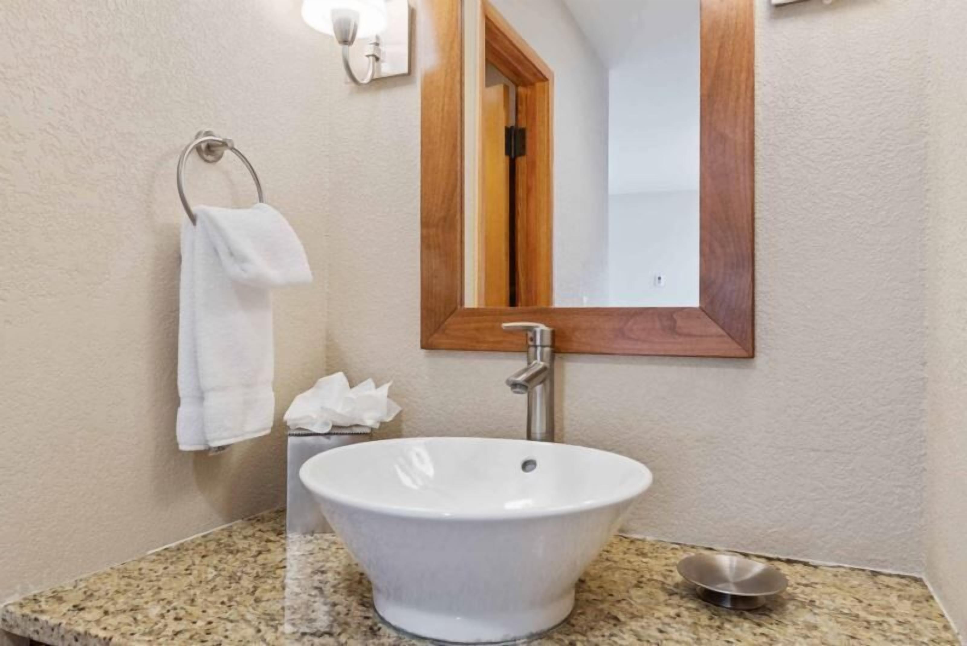 Un bagno con un lavandino bianco, uno specchio con cornice in legno, un asciugamano appeso a un anello metallico e una piccola ciotola metallica sul ripiano.