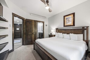 Une chambre d’hôtel avec un grand lit, une tête de lit en bois, un ventilateur de plafond, un tableau encadré au mur et une porte donnant sur une autre pièce.