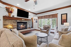 A living room with a fireplace, comfortable seating, a flat-screen TV, and a large window with a view of trees.