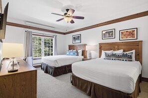 A hotel room with two beds, a ceiling fan, a desk with a lamp, and a window with curtains.
