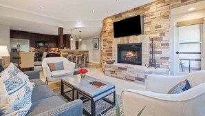 A modern living room with a stone fireplace, a kitchen in the background, and a dining area with bar stools. - The Charter at Beaver Creek A360 (Beaver Creek)