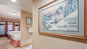 A framed picture of a winter sports scene in the Rockies, a bed, a window with curtains, and a door. - The Charter at Beaver Creek G055 (Beaver Creek)