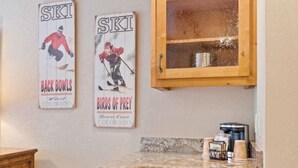 Une cuisine avec des meubles en bois, un plan de travail en granit et deux panneaux accrochés au mur, servant d’affiches pour promouvoir des événements liés au ski.