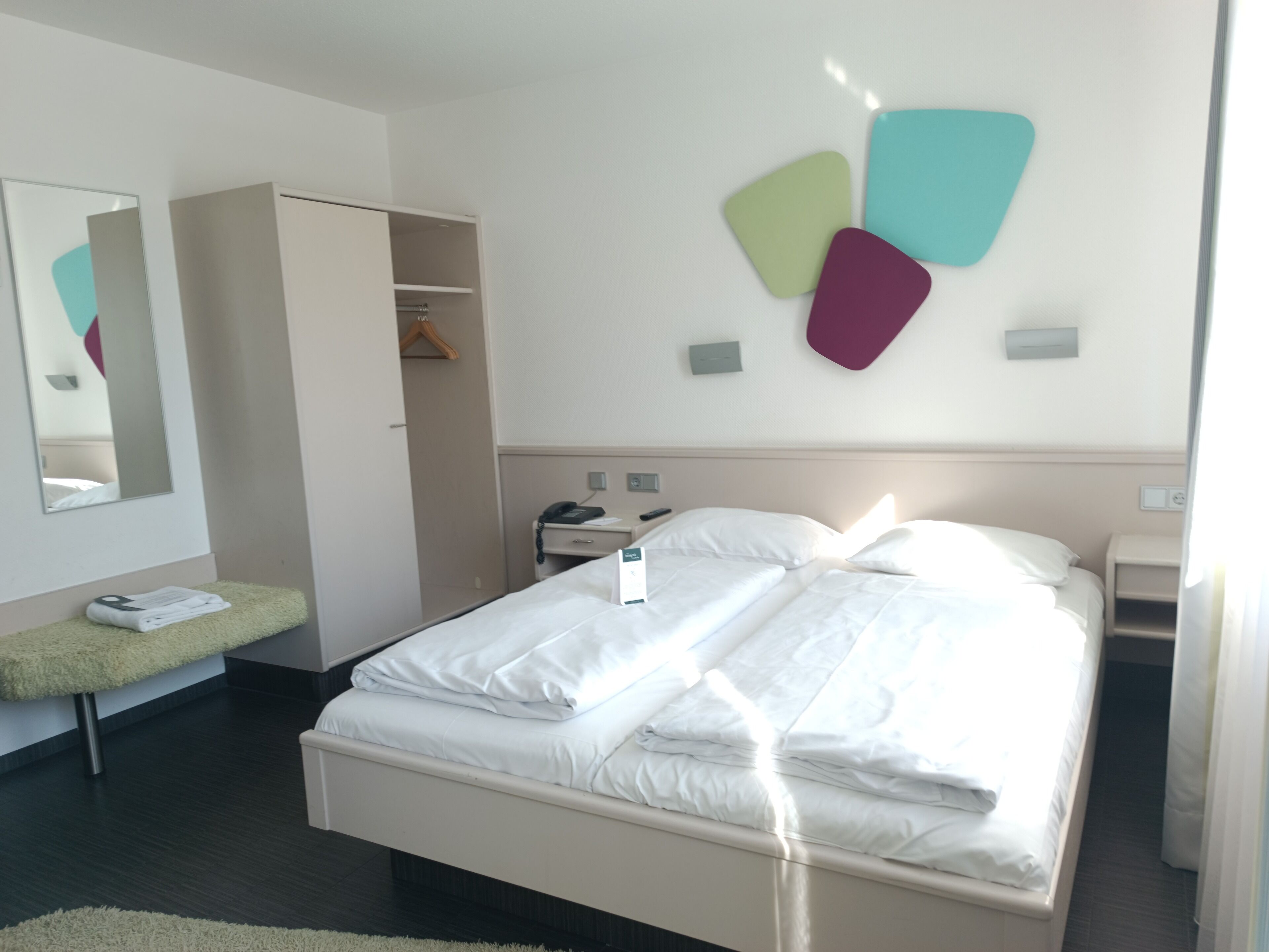 Moderni hotellihuone, jossa on siististi pedattu sänky, yöpöytä, jossa on puhelin, peili ja vaatekaappi.