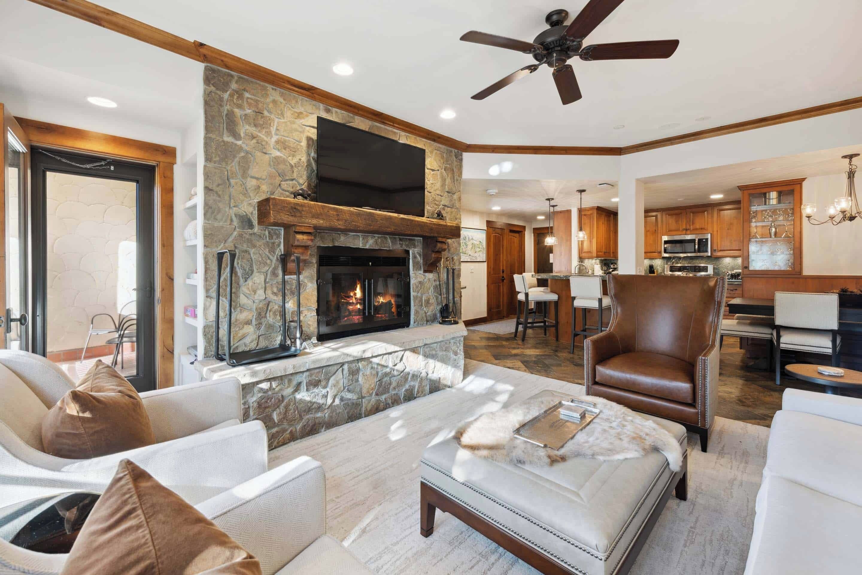 A spacious living room with a stone fireplace, comfortable seating, and a modern kitchen in the background.