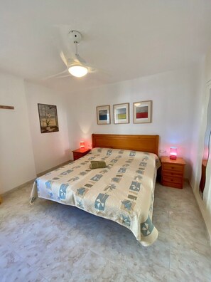 A bedroom with a bed, a ceiling fan, a nightstand with a lamp, and a wall-mounted shelf.