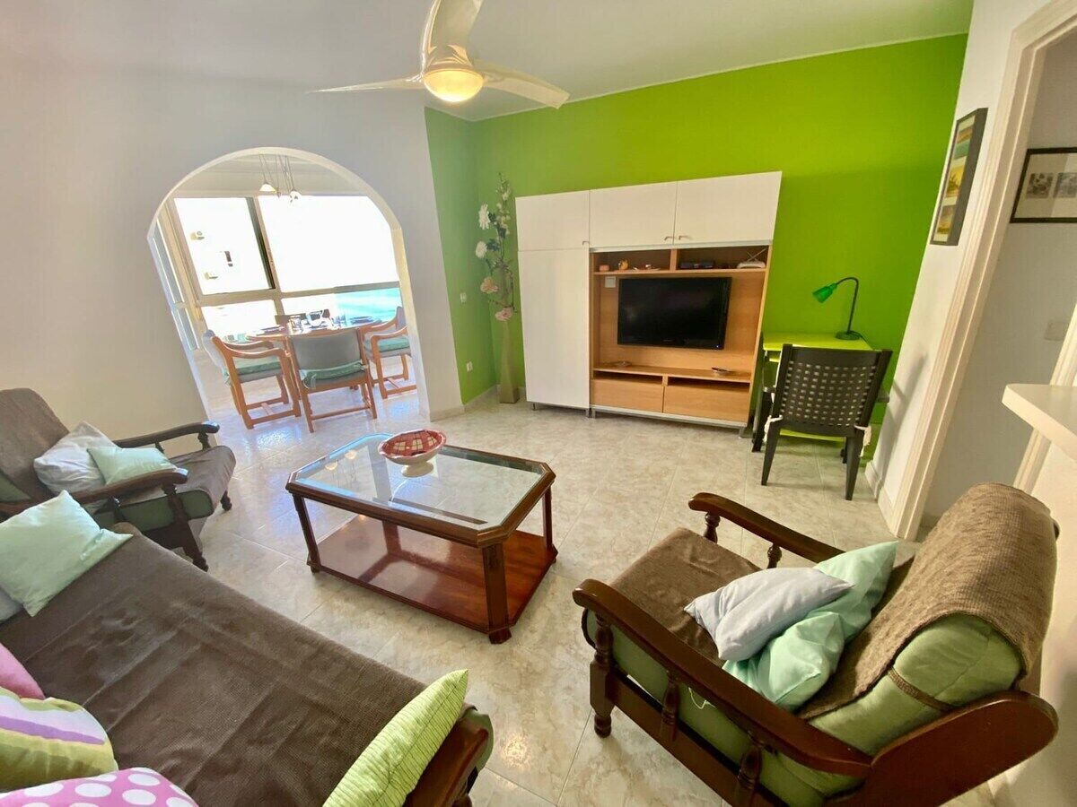 A living room with a TV, a glass coffee table, and a sofa. There is a green wall and a ceiling fan.