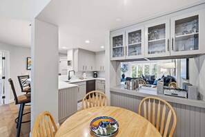 A modern kitchen with grey cabinets, a dining area with a wooden table and chairs, and a view of a living room with a sofa and a TV.