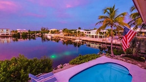 Una piscina con vistas a un puerto deportivo y palmeras, una bandera estadounidense y un cielo al atardecer.