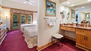 Une chambre d’hôtel avec deux lits, un grand miroir, une coiffeuse avec un lavabo et un ventilateur de plafond.