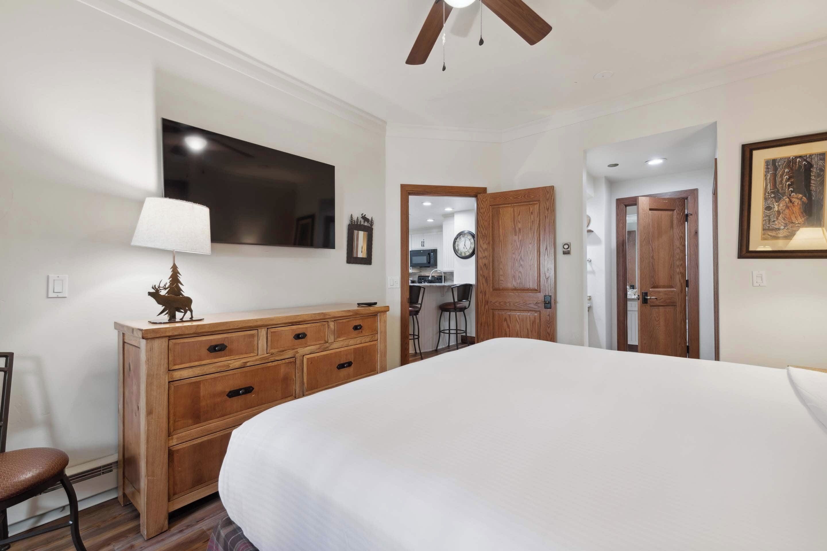 Un dormitorio con cama, cómoda de madera, lámpara y televisor montado en la pared.