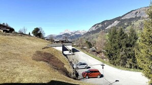 Une route de campagne avec une maison située sur une colline, une voiture garée sur le bord de la route et des montagnes en arrière-plan.