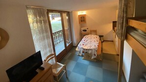 A compact room with a bed, a desk, and a chair. There is a window with curtains, a door, and a small table with a lamp.