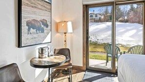 A hotel room with a bed, a table with chairs, a lamp, and a large window with a view of a snowy landscape.