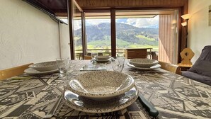 A dining area with a table set for four, a large bowl in the center, and a view of a mountainous landscape through a large window. - 300m from resort center, 1st floor, view ski slopes, balcony, parking, tv, ski locker, 35m² (Praz-sur-Arly)