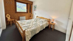 A bedroom with a wooden bed, a chair, a small table with a lamp, a window with a view of a forest, and a wall-mounted shelf. - 300m from resort center, 1st floor, view ski slopes, balcony, parking, tv, ski locker, 35m² (Praz-sur-Arly)