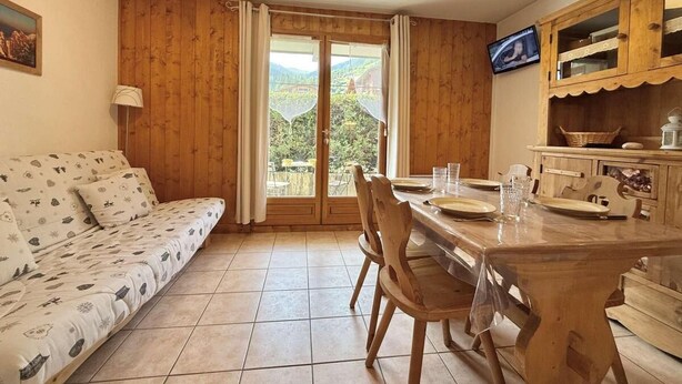 Une pièce chaleureuse avec un canapé en bois, une table à manger avec des chaises et une vue sur l’extérieur, visible à travers une porte vitrée.