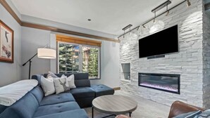 A modern living room with a stone wall, a flat-screen TV, a fireplace, and a large window with a view of greenery.