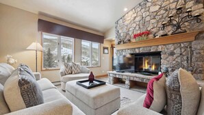 A cozy living room with a stone fireplace, comfortable seating, and a large window offering a view of snow-covered mountains. - The Charter at Beaver Creek D440 (Beaver Creek)