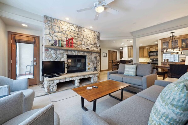 A living room with a stone fireplace, a television, and a coffee table. There is a sofa and a dining area in the background. - The Charter at Beaver Creek D340 (Beaver Creek)