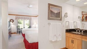 Une chambre d’hôtel avec un lit, un bureau, une chaise, une fenêtre donnant sur un paysage agréable, une salle de bain avec un lavabo et un miroir, et des serviettes accrochées au mur.
