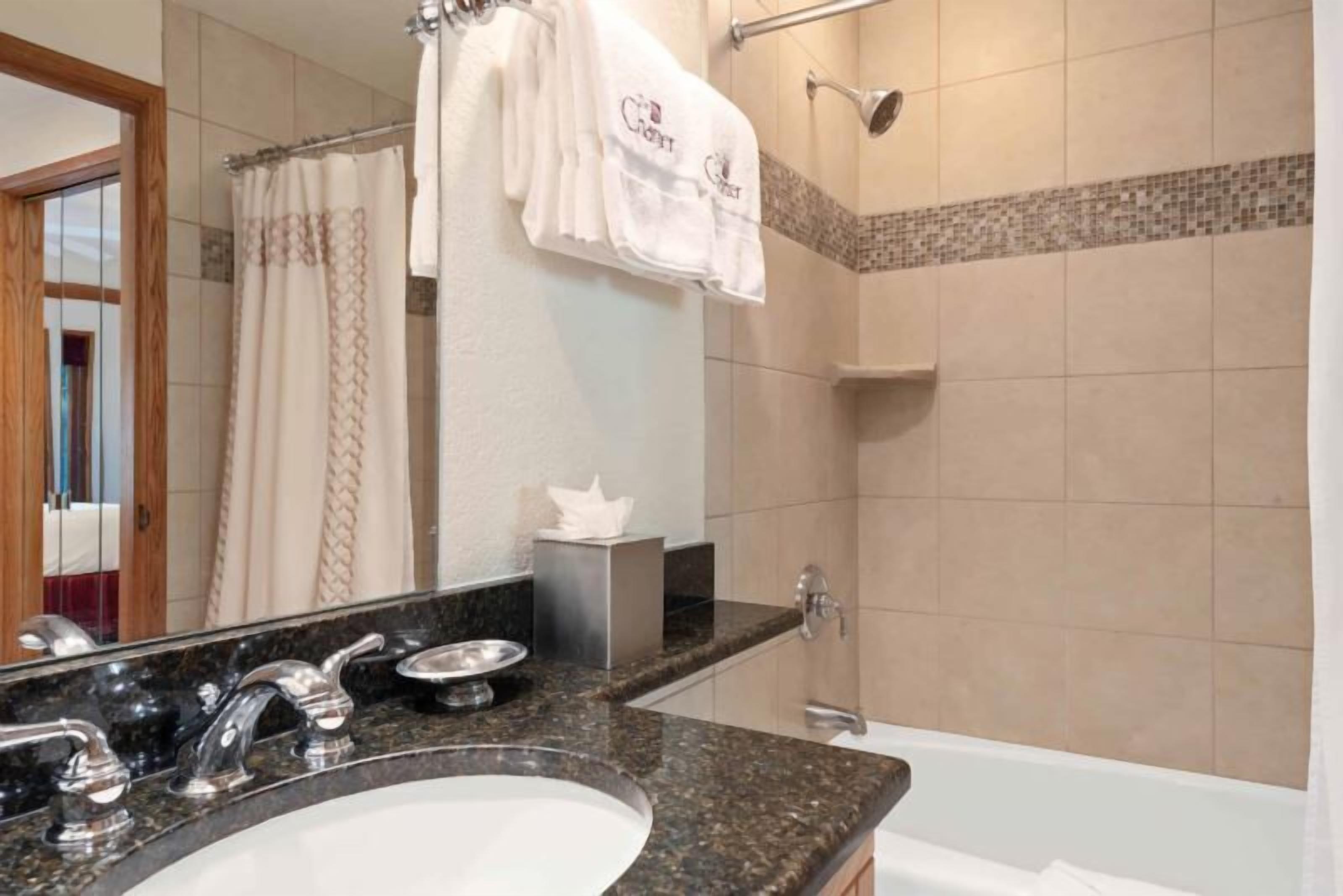 Un baño con lavamanos de granito, dos grifos, un espejo, toallero con toallas blancas, cortina de ducha y zona de ducha con azulejos.