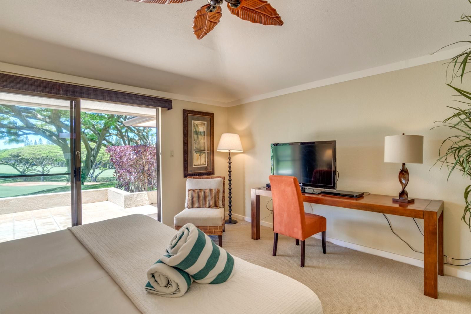 A hotel room with a bed, a desk with a TV, a chair, a lamp, and a view of a garden.