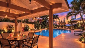 A covered outdoor pool area with lounge chairs, a dining table with drinks, and tropical plants.