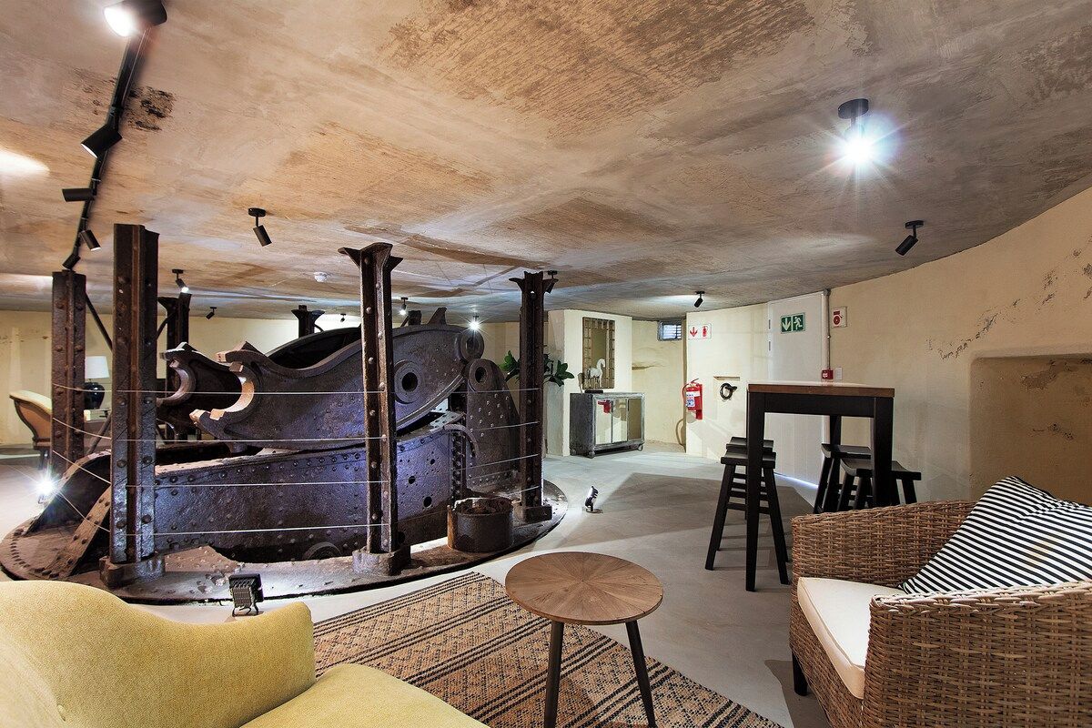 A room with a large industrial machine, a dining area with a table and chairs, and a wicker sofa.