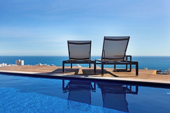 Un área junto a la piscina con dos sillas mirando al océano, una terraza de madera y una vista despejada del mar.