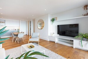 A modern living room with a flat-screen TV, a dining area with chairs, and a kitchen in the background.