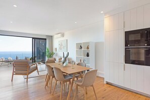 Eine moderne Küche mit weißen Schränken, ein Essbereich mit Holztisch und Stühlen sowie Meerblick durch große Fenster.