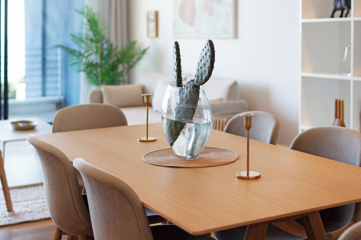A dining table with a clear glass vase containing two cacti, surrounded by chairs and a modern lamp.