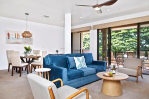 A modern living room with a blue sofa, a dining table, and chairs. There is a large window with a view of trees. - Kapalua Golf Villas | Golf View 2 BR | Car Incl. w/ 6+ Nights | KGV-14V3 by KBM (Lahaina)