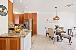 A modern kitchen with wooden cabinets, a stainless steel refrigerator, granite countertops, and a dining area with a round table and chairs. - Kapalua Golf Villas | Golf View 2 BR | Car Incl. w/ 6+ Nights | KGV-14V3 by KBM (Lahaina)