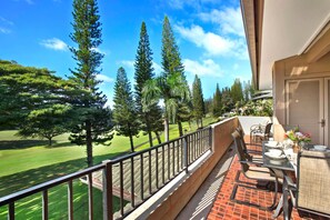 A balcony with a view of a golf course, a dining area with outdoor furniture, and a closed shutter door. - Kapalua Golf Villas | Golf View 2 BR | Car Incl. w/ 6+ Nights | KGV-14V3 by KBM (Lahaina)
