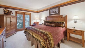 Une chambre à coucher avec un grand lit, une tête de lit en bois, deux lampes de chevet, une table de chevet, une commode et une fenêtre donnant sur les arbres.