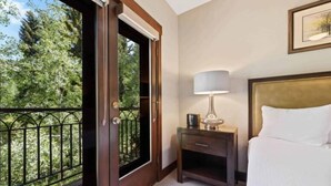 A hotel room with a bed, a nightstand, a lamp, and a window with a view of trees. - The Charter at Beaver Creek C230 (Beaver Creek)