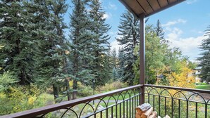 Ein Balkon mit Metallgeländer, Holzstapel und Blick auf ein bewaldetes Gebiet mit hohen Nadelbäumen und ein Gebäude im Hintergrund.