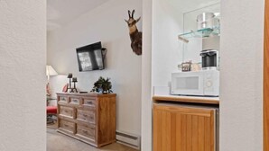 A kitchen with a microwave, a wooden cabinet, a wall-mounted shelf with kitchen items, and a mounted animal head decoration.
