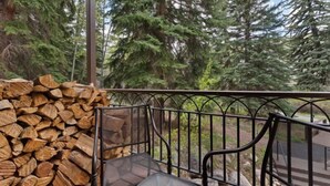 Un balcon avec une pile de bois de chauffage, des chaises en métal et une vue sur de grands arbres à feuilles persistantes.