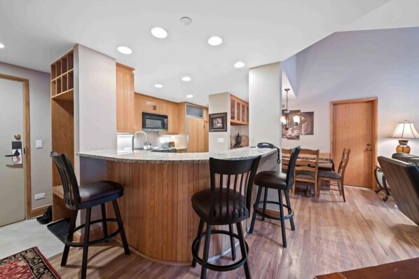 Cocina con gabinetes de madera, una isla central con taburetes de bar, una zona de comedor con mesa y sillas, e iluminación empotrada en el techo.