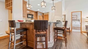 A modern kitchen with wooden cabinets, a central island with stools, pendant lighting, and a visible microwave.