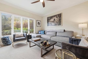 A living room with a grey sofa, a coffee table, and a chair. There is a painting on the wall and a window with a view of trees. - The Charter B310 - 2 Bedroom (Beaver Creek)