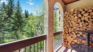 Un balcon donnant sur une forêt de conifères, une pile de bois de chauffage et une table avec des chaises.