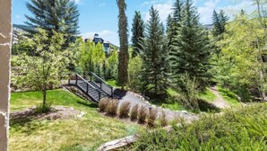 Un pont en bois enjambe un ruisseau dans un parc où se dressent de grands arbres, et on aperçoit un bâtiment au loin.
