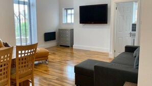 A modern living room with a flat-screen TV, wooden flooring, a grey sofa, and a dining area with a wooden table and chairs. - Inviting 1-bed Apartment in Lytham Saint Annes (Lytham St. Anne's)